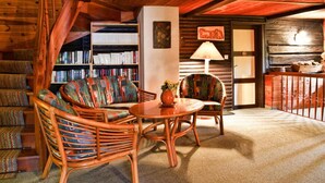 A cozy log cabin interior with a wooden staircase, a bookshelf, a dining area with a table and chairs, and a lamp.