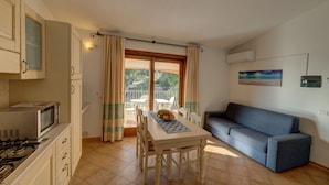 A compact kitchen with a microwave, stove, and cabinets. There's a dining table with chairs and a fruit bowl. A sofa is against the wall, and a window with curtains is visible. - Apartment in Badus With Communal Pool (Badesi)