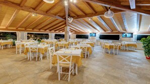 A spacious indoor dining area with multiple tables and chairs, wooden beams on the ceiling, and ceiling fans. - Apartment in Badus With Communal Pool (Badesi)