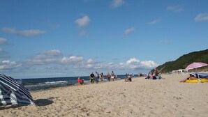 En sandstrand med folk som promenerar och sitter, ett randigt parasoll och en kulle i bakgrunden.