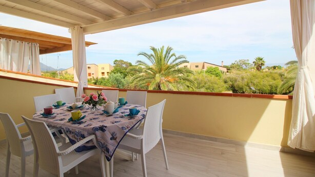 A balcony with a table set for two, white wicker chairs, and a view of greenery and buildings in the distance. - Apartment on Costa Rei With Stunning Beaches (Muravera)