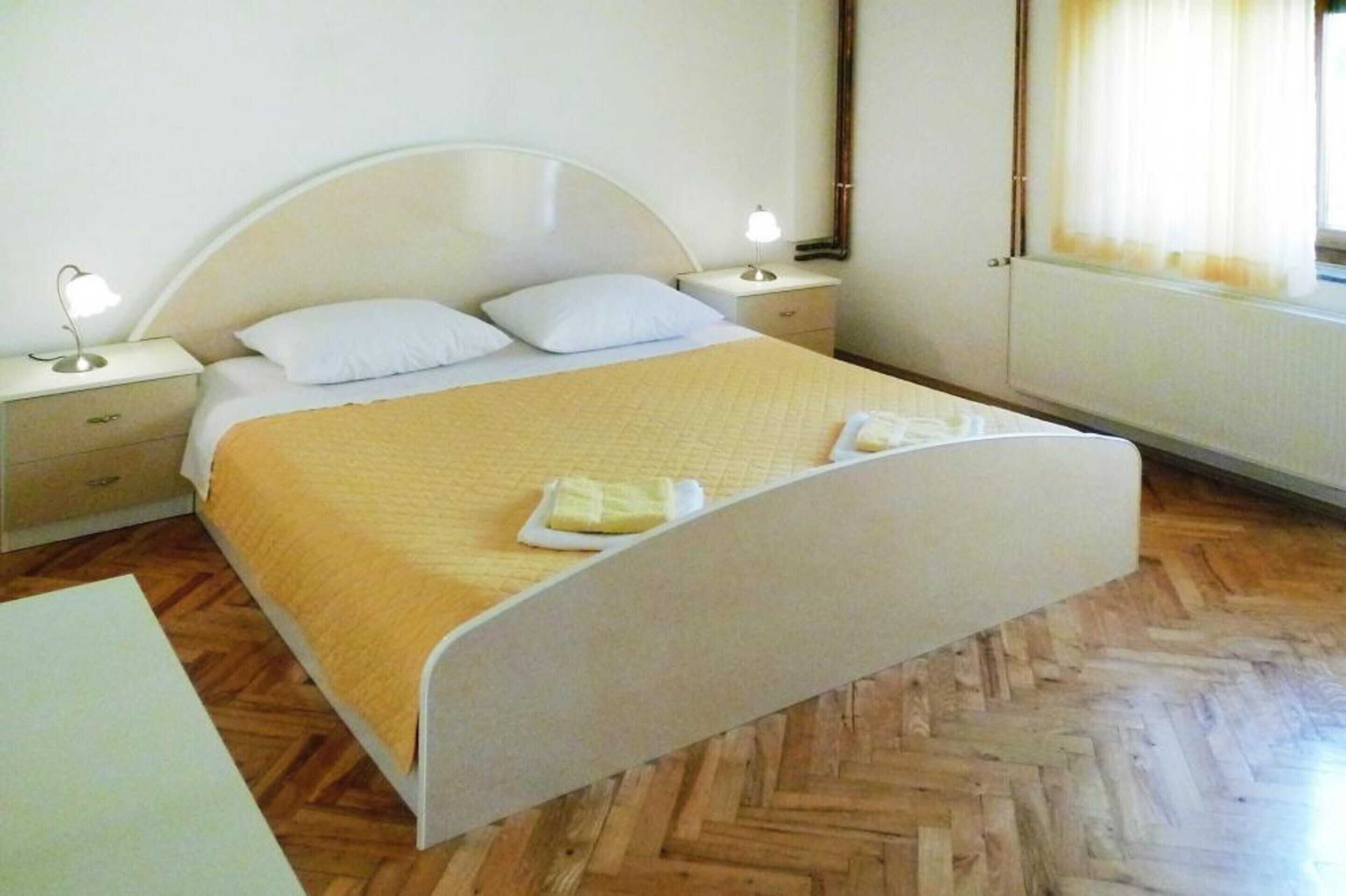 A bed with a headboard, two bedside tables with lamps, a wooden herringbone floor, and a window with curtains.