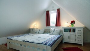 Une chambre à coucher avec un lit en bois, des tables de chevet blanches, une table de nuit, un tapis rouge, une fenêtre avec des rideaux rouges et une petite armoire contenant des livres et une plante.