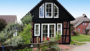Une maison en noir et blanc, avec un toit de chaume, une clôture en bois portant le numéro 36, et un jardin agrémenté de plantes et de fleurs.