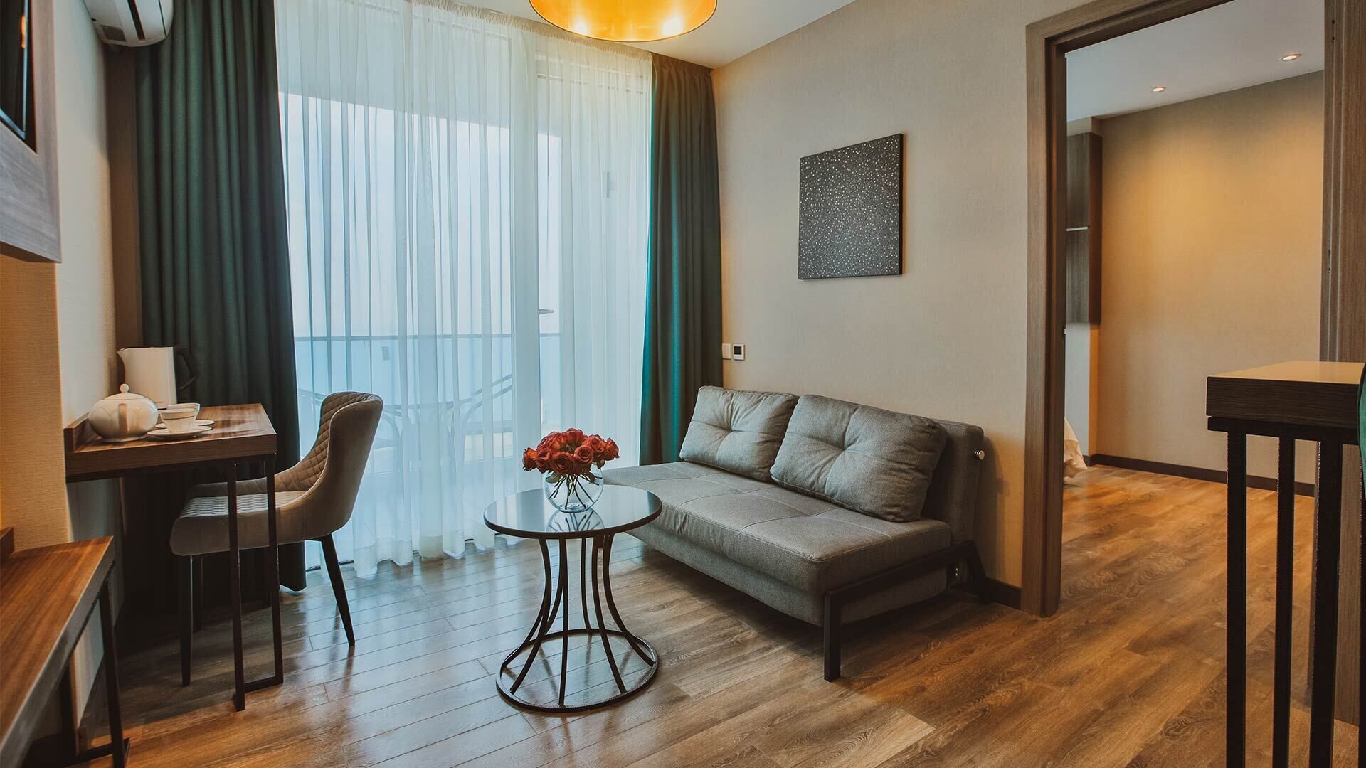 Ein modernes Hotelzimmer mit einer Sitzecke, einem runden Tisch mit Blumenvase, einem Essbereich mit Stuhl und Tisch sowie einem großen Fenster mit Vorhängen.