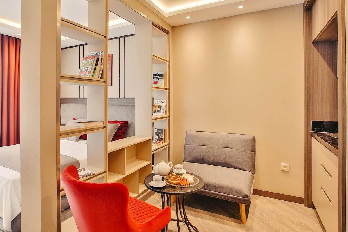 Ein Hotelzimmer mit einem Bett, einem roten Sessel, einem kleinen Tisch mit Teekanne und Tassen sowie einem Bücherregal mit diversen Gegenständen.