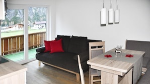 Un salón moderno con un sofá gris oscuro, cojines rojos, una mesa de comedor de madera con sillas grises, y vistas a una terraza de madera y montañas a través de amplios ventanales.