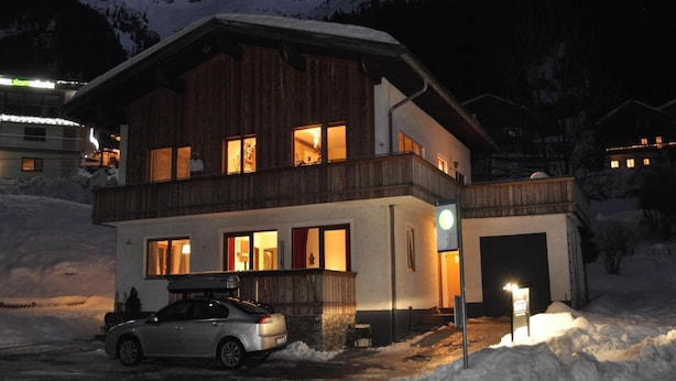 Un chalet de madera de dos pisos con un coche estacionado frente, rodeado de montañas nevadas.