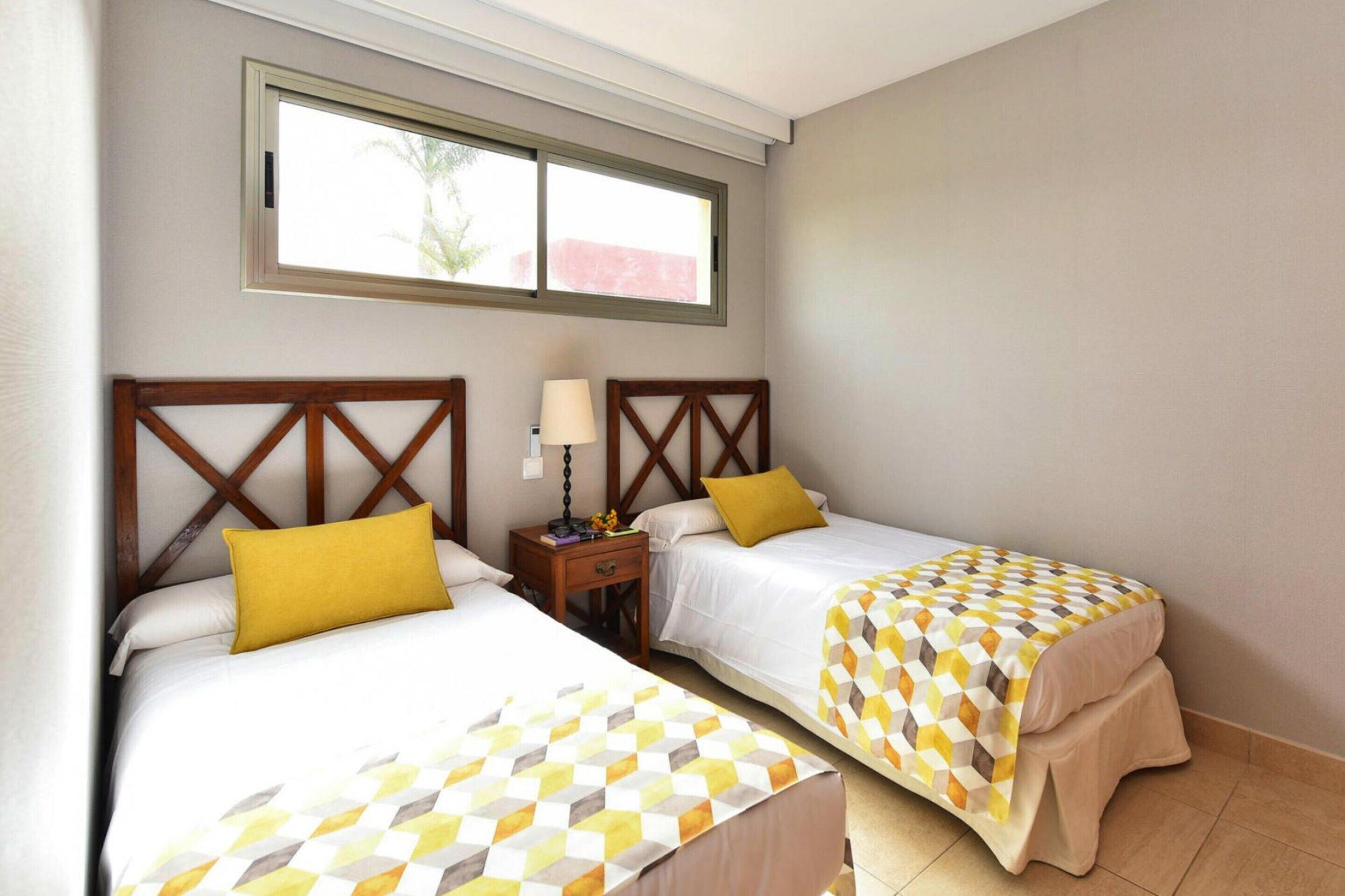 Une chambre d’hôtel avec deux lits, chacun doté d’une tête de lit en bois et d’une couvre-lit à motifs. Il y a une table de chevet avec une lampe entre les deux lits, et une fenêtre au-dessus des lits.