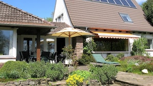 A two-story house with a sloped roof, a covered patio with outdoor furniture, and a garden with various plants and a yellow flower bed. - Ferienhaus fur bis zu 6 Personen, Am Wung in Husum (Husum)