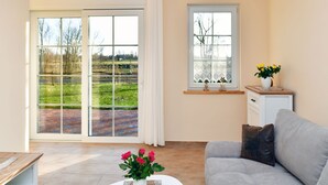 Uma sala de estar moderna com um sofá cinzento, um armário branco pequeno e uma janela com vista para uma paisagem verde.
