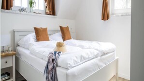 A bedroom with a white bed, brown curtains, a window, and a wooden floor. - Ferienwohnung, Zingst (Zingst)