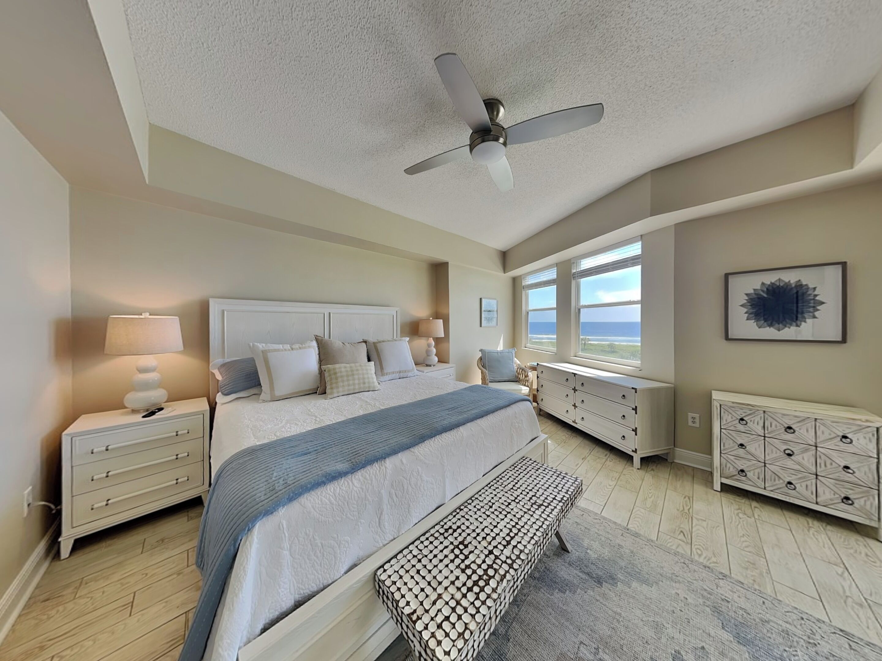 A bedroom with a large bed, a ceiling fan, a view of the ocean, and a dresser with drawers.