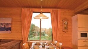 Interior de uma cabana de madeira com uma mesa de jantar posta para quatro pessoas, um relógio na parede e uma janela com cortinas.