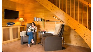 Interior acolhedor de uma cabana com paredes de madeira, uma televisão, uma estante de livros e duas poltronas reclináveis.