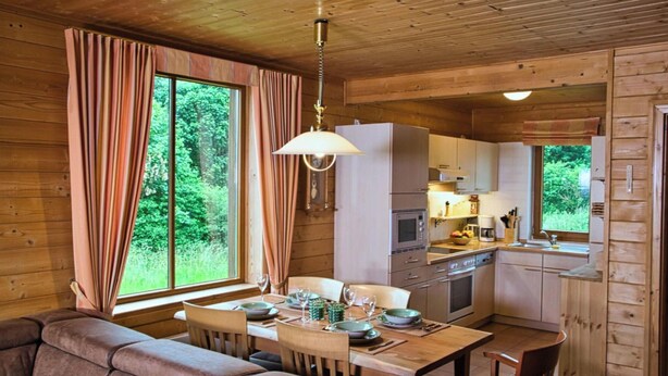 Interior de uma cabana de madeira com área de jantar, cozinha e janelas amplas.