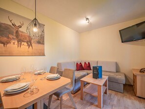 Una sala moderna con una mesa de comedor preparada para cuatro personas, un sofá gris con cojines rojos, un televisor de pantalla plana montado en la pared y una lámpara de techo.