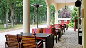 An outdoor patio with wooden tables and chairs, stone flooring, and a view of trees and a building in the background. - Ferienpark Markgrafenheide (Rostock)