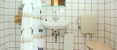 Ein Badezimmer mit weißem Waschbecken, Toilette und Dusche. Über dem Waschbecken befindet sich ein Spiegel und eine Handtuch an der Wand.