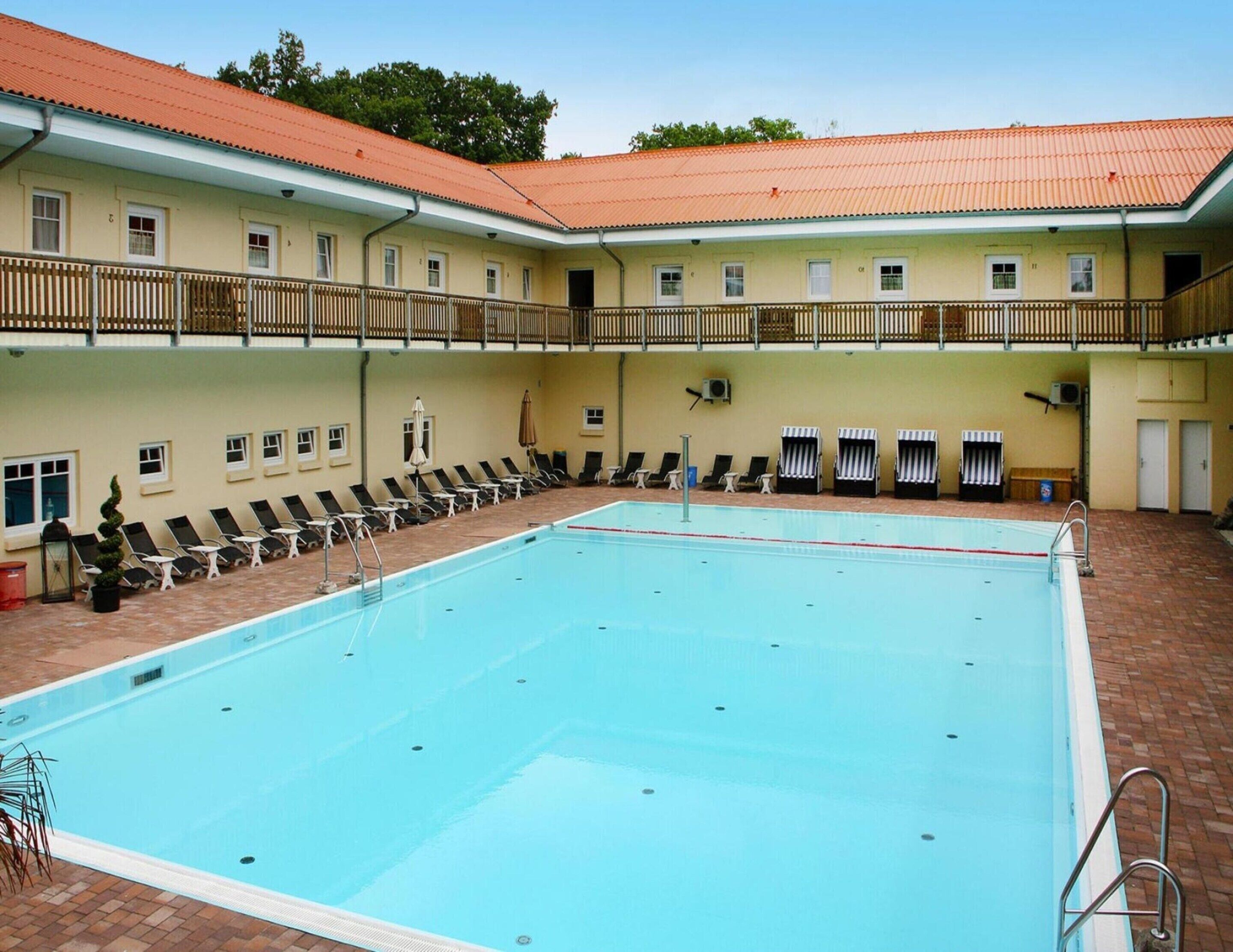 A swimming pool with lounge chairs and umbrellas, a covered seating area, and a building with a red-tiled roof.