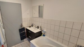 Et badeværelse med badekar, vask og spejl. Der er håndklæder hængende på en håndklæderække og en dør til et andet rum.