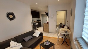 En kompakt opholdsstue med en mørk sofa, et lille rundt bord og et spiseområde med et hvidt bord og stole. Der kan ses et køkken i baggrunden, og en radiator er placeret nær vinduet.