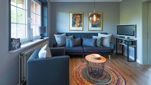 A modern living room with a dark sofa, a coffee table, and a television. There are two framed artworks on the wall and a window with books on the sill.