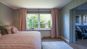 A bedroom with a large bed, a window with curtains, a radiator, and a view of greenery outside.