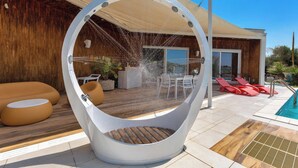 Area piscina esterna moderna con una struttura di sedute a forma circolare, un pontile in legno e poltrone da relax rosse.