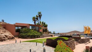 A modern building with a stone-paved entrance, surrounded by greenery and palm trees.