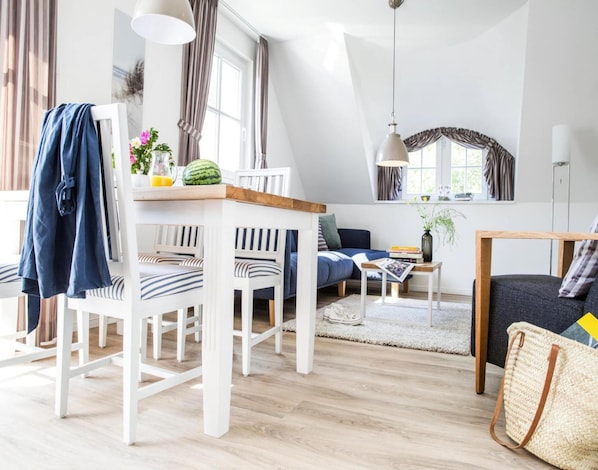 A modern living room with a dining area, featuring white furniture, a wooden table, and a large window with curtains. - Wohnung in Zingst Nahe Ostseestrand (Zingst)