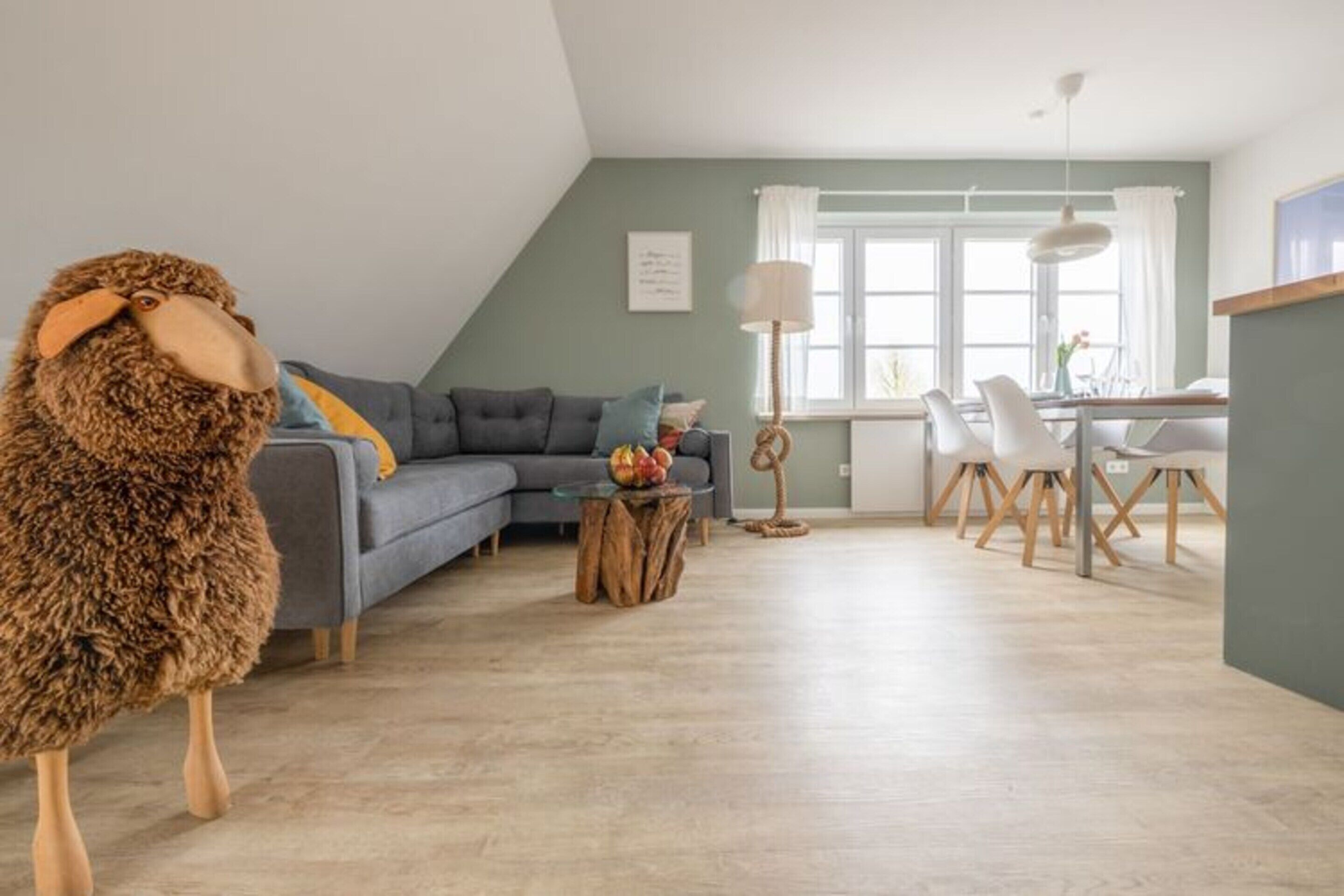 Ein modernes Wohnzimmer mit einem grauen Sofa, einem Esstisch mit Stühlen und einer großen Stofftierfigur.