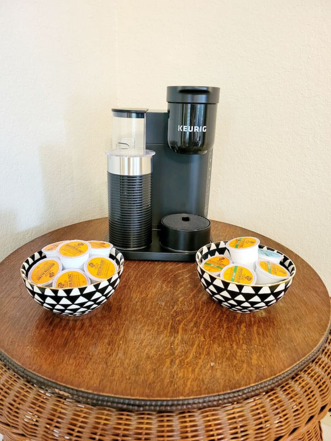 Een Keurig koffiezetapparaat op een ronde houten tafel, met twee gemêleerde kommen gevuld met koffiekapsules.
