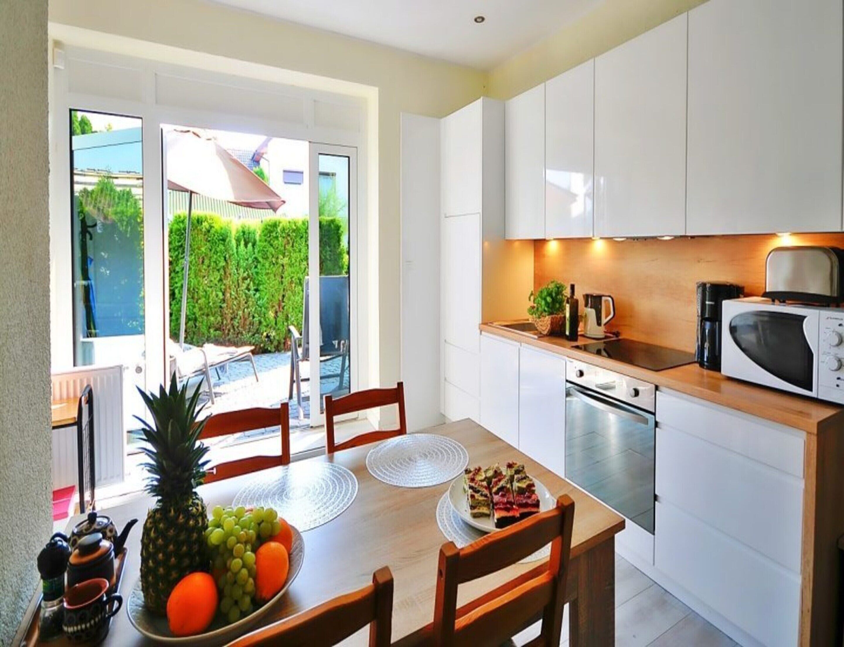 A compact kitchen with white cabinets, a microwave, and a coffee maker. A dining table with chairs and a fruit bowl is visible.