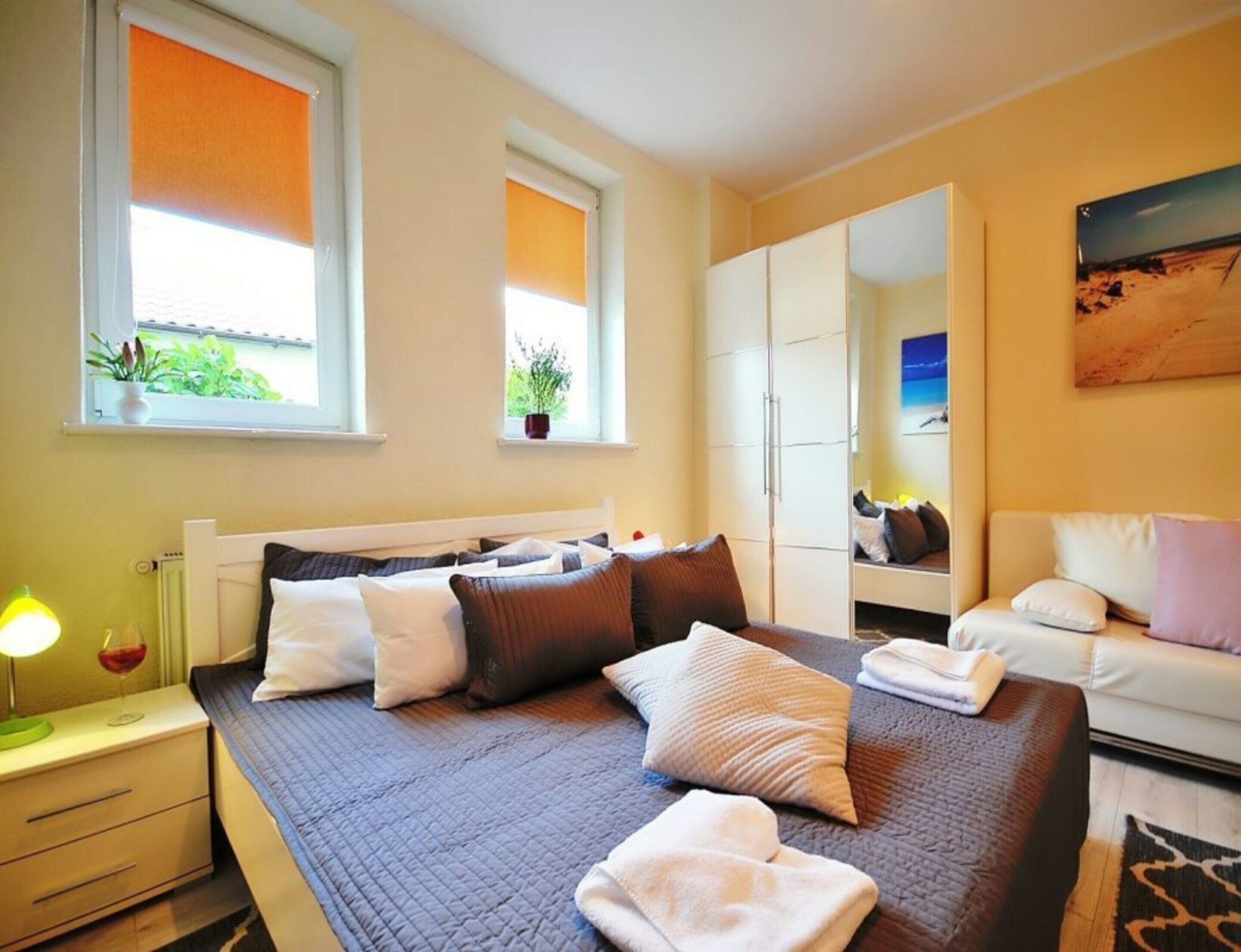 A bedroom with a bed, bedside table, lamp, windows with blinds, a wardrobe, and a painting on the wall.