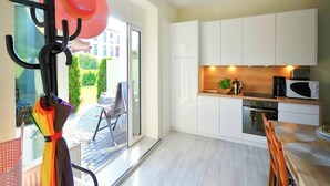 Eine moderne Küche mit weißen Schränken, einer Mikrowelle und einer Kaffeemaschine. Es gibt eine Essecke mit Tisch und Stühlen, und eine Glastür, die auf eine Terrasse mit einem Stuhl und Tisch führt.