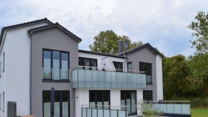 Moderni, kaksikerroksinen talo, jossa on parveke, suuret ikkunat ja pieni puutarha-alue.