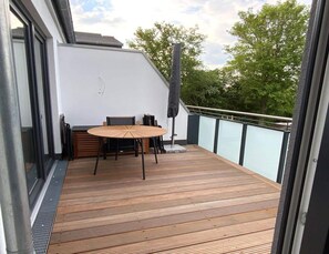 A wooden deck with a table and chairs, a tall umbrella, and a glass balustrade. - Ferienwohnung Inselresort James Cook auf Fehmarn (Fehmarn)