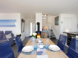 A dining area with a wooden table set for four, flanked by blue chairs, a white sofa, and a painting of a beach scene. A fireplace with a vase of white flowers is in the background.