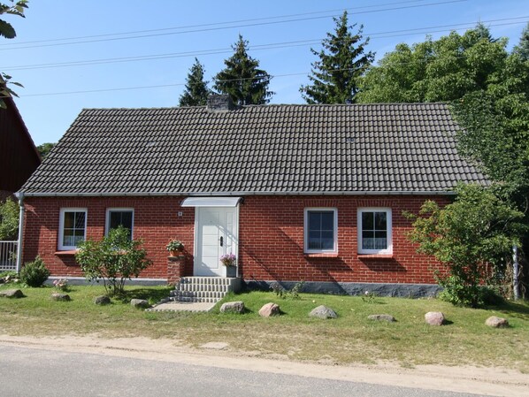 Une maison en briques rouges, avec un toit en tuiles, une porte d’entrée et deux fenêtres. On peut voir des rochers et un petit arbre devant la maison.