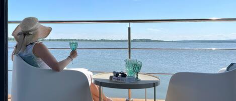 A person seated at a table on a deck with a view of a body of water and land in the distance.