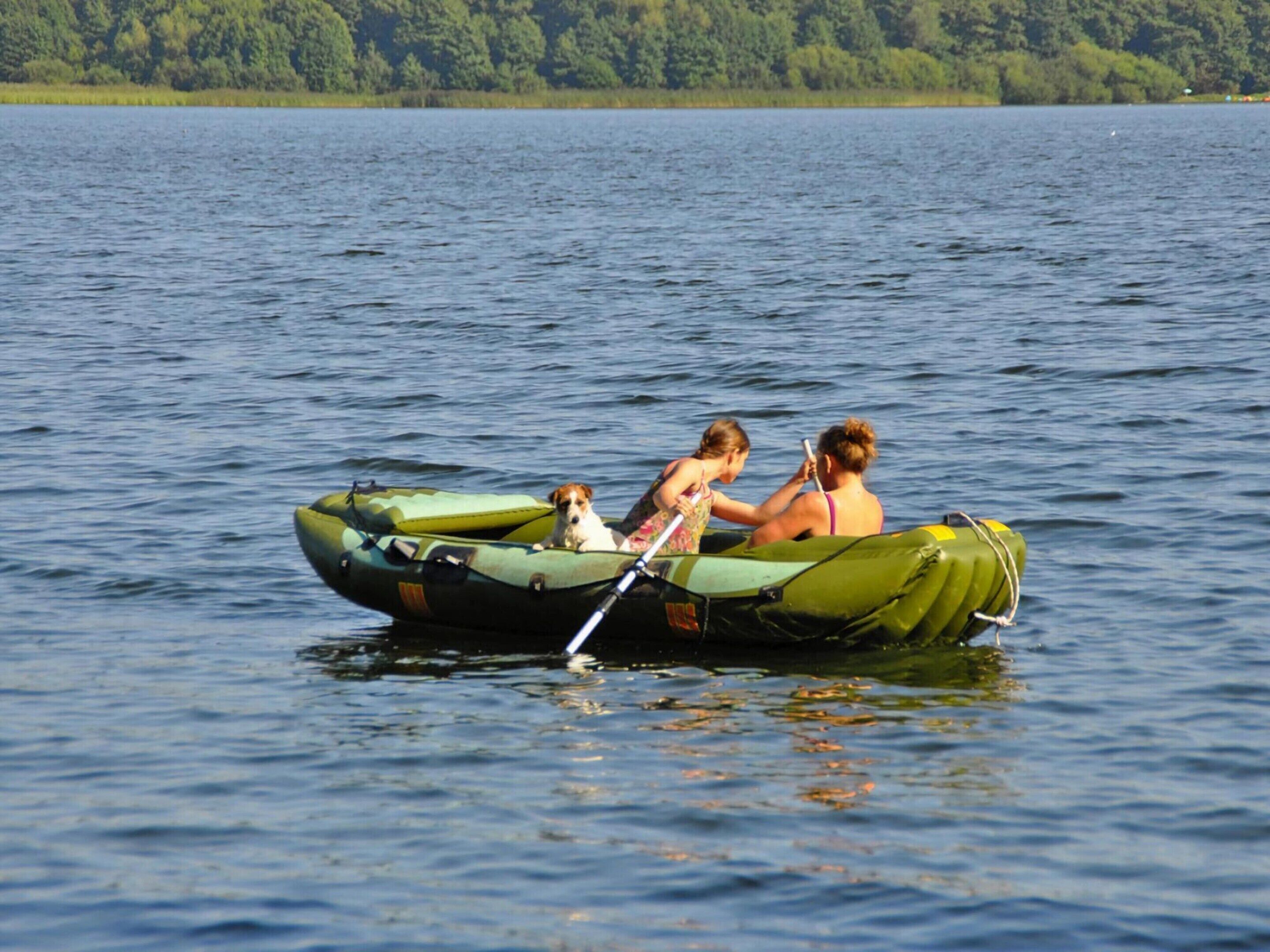 A green inflatable boat on calm water with two people and a dog.