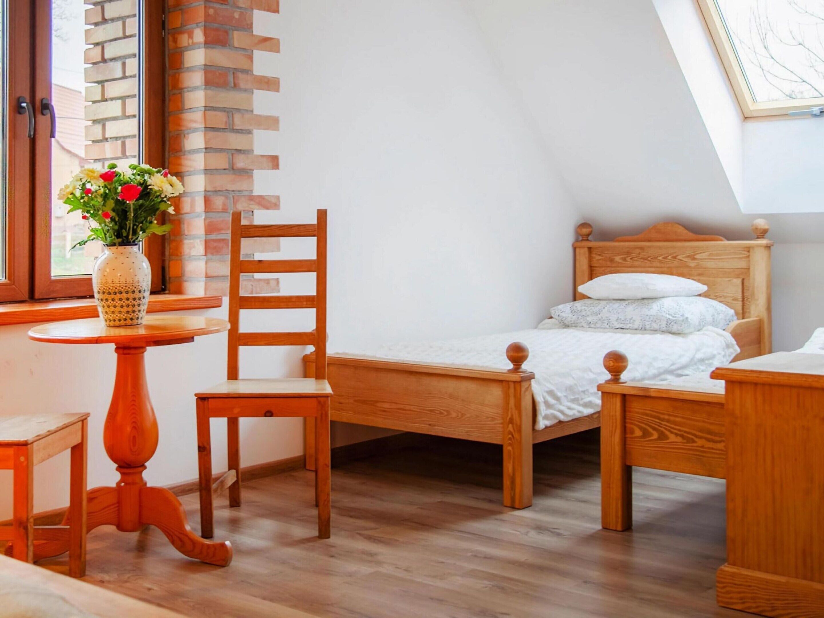A bedroom with a wooden bed, a wooden chair, a small wooden table, and a window with a brick wall.