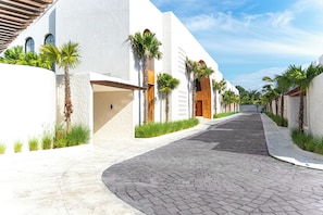 A paved pathway flanked by white buildings with wooden doors and palm trees. - Nila Residence (Canggu)
