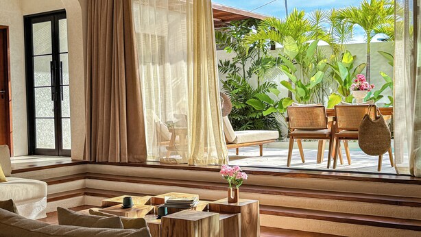 A living room with a sofa, wooden coffee table, and chairs. There is a window with sheer curtains and a view of greenery outside. - Nila Residence (Canggu)