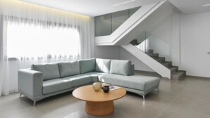Una sala de estar moderna con un sofá seccional gris claro, una mesa de centro de madera redonda y una escalera con barandillas de vidrio.