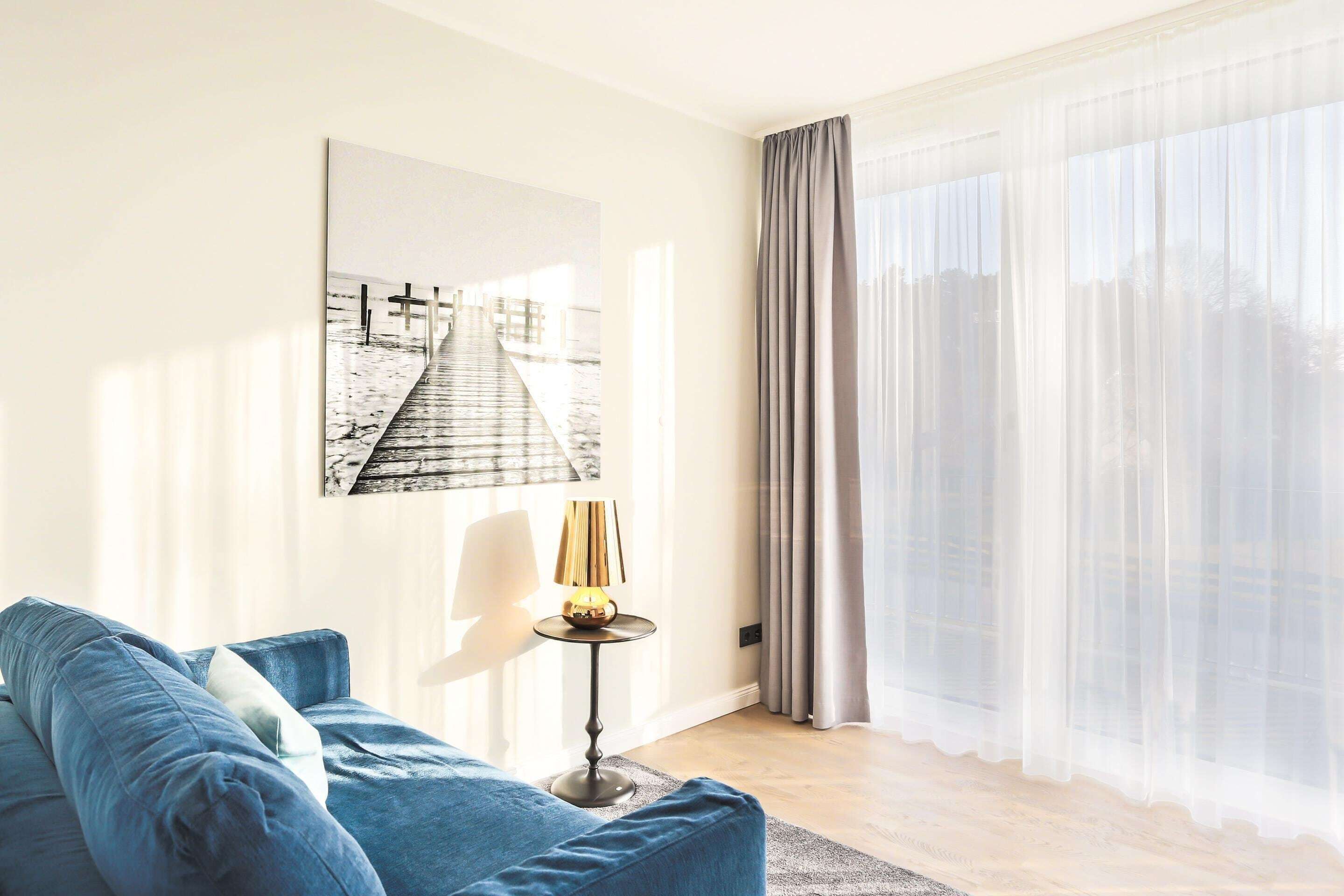 A modern living room with a blue sofa, a small round table with a lamp, a large window with sheer curtains, and a framed artwork on the wall.