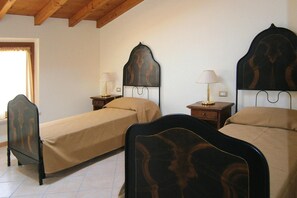 A room with three single beds, each with a wooden headboard and a beige bedspread. There are bedside tables with lamps and a small drawer. The room has a wooden ceiling and a window with a curtain. - Apartment in Lazise With Parking Space (Lazise)