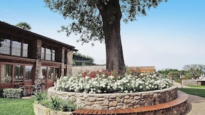 A building with a stone facade, a large tree, and a flower bed with white and red flowers. - Apartment in Lazise With Parking Space (Lazise)