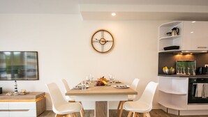 A modern dining area with a wooden table, white chairs, and a wall-mounted clock. - Ferienhaus in Damp am Ostseestrand (Damp)